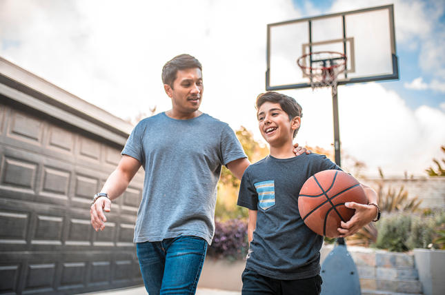 A father and son stand smiling in their driveway. The father has his arm on the son's shoulder. The son is holding a basketball, and a basketball hoop is shown in the background.